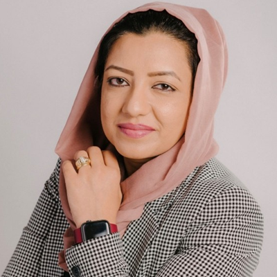 Headshot of Farkhanda Ehssan wearing a black and white blazer and pink headscarf against a grey background
